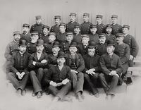 Ansatte ved Trondheim straffeanstalt. Fengselsdirektør Daae, som Lars Erikssons skulle besøke i 1894, er sittende foran. Betjent John Herstad, som blødde ihjel få minutter etter Erikssons knivstikk i 1894, sitter helt til høyre i nederste rad av menn med uniformslue. Helt til venstre i samme rekke sitter, betjent Wøien, og nr. 3 fra venstre i bakerste rekke er betjent Husby, som begge ble knivstukket av Lars Eriksson, men overlevde. Underinspektør Eriksen, som beordret visitasjon av Lars Eriksson, sitter som nr 4 fra venstre i samme rekke som Herstad. Foto: Justismuseet