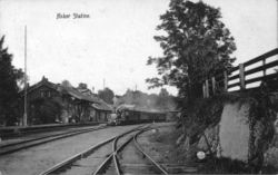 Asker stasjon sett fra sør med den opprinnelige stasjonsbygningen, tegnet av G. A. Bull, fra 1872. Et tog fra Kristiania på vei til Drammen står ved midtplattformen med dampen oppe. Foto fra J. Michelet: En hilsen fra Asker. Foto: Ukjent