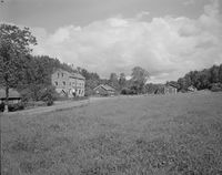 Lommedalsveien sett mot Bærums Verk. I venstre bildekant, nr. 180, arbeidsboligen Island, den to etasjes murbygningen er nr. 182, småskolen fra 1900, fra 1936 Bærums Verk Handelsforening, den 1 ½ etasjes trebygningen er nr. 184, oppført på tomten til den tidligere storskolen fra 1881, etter at denne brant ned, på den andre siden av veien, til høyre i bildet er nr. 189, arbeiderboligen Stortinget. Jordet i forkant av bildet heter Kristiansjordet og er fortsatt i drift. Foto: R. Andersen/Nasjonalbiblioteket (1956).