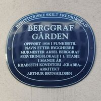 Blå oval byhistorisk plakett på vegg i Fredrikstad. Skiltet forteller om Berggrafgården, oppført i 1938 i funkisstil, oppkalt etter byggmester Aksel Berggraf. Bygget hadde serveringslokale i 1. etasje i mange år, blant annet konditoriet «Krabba». Arkitekt var Arthur Brynhildsen.