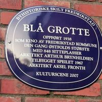 Blå oval byhistorisk plakett på murvegg i Fredrikstad. Skiltet forteller om Blå Grotte, oppført i 1938 som kino av Fredrikstad kommune, den gang Østfolds største med 848 sitteplasser. Arkitekt Arthur Brynhildsen. Bygget ut med Speilet i 1967 av arkitekt Aksel Fronth og tatt i bruk som kulturscene i 2007.