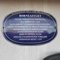Blå oval byhistorisk plakett på vegg i Fredrikstad. Skiltet forteller om Børneasyllet i Vestre Fredrikstad, opprettet i 1864 som daghjem for barn av fattige eller enslige arbeidende foreldre. Lå ved hjørnet av Nygaards gaten og Asylgaten. Bygningen ble revet i 1934.