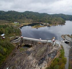 Dammen sett fra lufta, den var opprinnelig en nåledam, men ble i 2012 blendet igjen med bruddstein som tilsvarende den originale steinen. Flomlukene til høyre. Foto: Dag Endre Opedal/Norsk Vasskraft- og Industristadmuseum (2016).