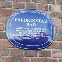 Blå oval byhistorisk plakett på murvegg i Fredrikstad. Skiltet forteller om Fredrikstad Bad, åpnet som folkebad i 1888 med badstue og karbad, drevet som privat aksjeselskap og senere av kommunen fra 1936, nedlagt i 1971.