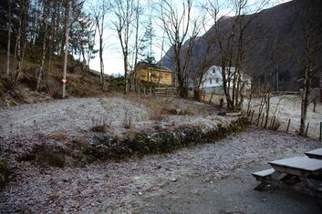 Ein mosegrodd mur kutter av ei helling i terrenget. Under muren er ein gruslagd plass kor to benkar er synlege i det eine hjørnet av bildet. Gras og andre småplantar veks over og rundt sjølve muren. Bildet er tatt på seinhøsten, og det ligg lauv på bakken. Eit lag rimfrost dekkar heile området.