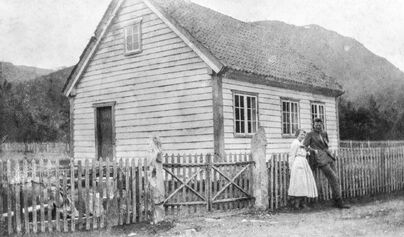 Svart-kvitt foto av hus med gjerde rundt. To personar står lent mot gjerdet.