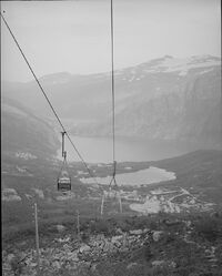 Taubane i tettstedet Glomfjord med Litlglomvatnet og Glomfjorden i bakgrunnen. Glomfjord kraftverk kan skimtes innerst i fjorden til venstre. Dette er det også en pendelbane for offentlig persontransport, her bygd som stolheis. Åpne stolheiser skal normalt ikke være mer enn femten meter over bakken.
