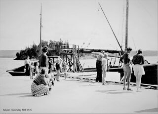 Islasting på brygga på Ramton. Isen ble tatt ut av isstablene og vasket før den ble sendt ut på brygga og lastet om bord i lastejakta. Midt på bildet blir en isblokk skjøvet fram på ledere av plankebord som var lagt fram til kanten av brygga. Foto fra 1953-1954. Fotograf ukjent.