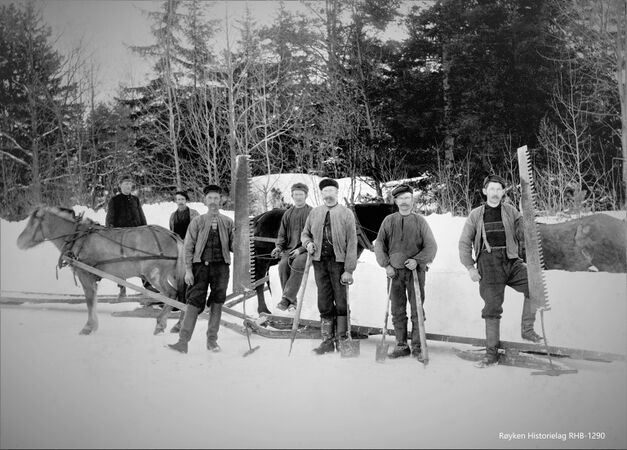 ISARBEIDERE PÅ BÅRSRUDTJERNET CA 1903-1905.Isskjærere og kjørere på Bårsrudtjernet (Nærsnes). Hesten «Blakken» er spent foran en isslådd (lang slede). Isskjærere viser fram redskapene; issager, ishakker (til å løfte/manøvrere isblokkene) og isøkser. Martin Skryseth ved «Blakken», Kristian Skryseth sitter på islasten. Foran Islasten står fire isskjærere, trolig fra Konnerud. (Foto fra Røyken Historielag, fotograf Johan K A Foss)