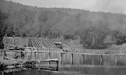 ISSTABEL PÅ FLATASTRANDA 1890-TALLET. Den korte brygga nærmest går ut fra det som i dag er Propsen. Den neste er brygga (isrenna) fra Istsablen på Mellomstranda og innenfor den ligger isrenna fra Flatstranda. I bakkant av isstabelen ser vi ei isrenne som førte is som kom inn fra Scwartzedammen, Til høyre på stabelen ser vi at det var tilrettelagt for at isen tas ut fra flere forskjellige høyder. Utsnitt av større fotografi. (Foto fra AS Søndre Nærsnes)
