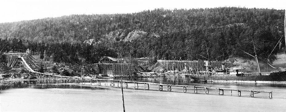 Dette bildet fra 1894-1898 viser hvordan veggene på isstablene ble støttet opp og den lange isrenna for lasting om bord i skip. De rentspylte isblokkene ble sendt ut på isbrygga og skled ut til båter som heiste blokkene om bord for videre transport. Til høyere sees et permanent ishus med saltak. Disse måtte etter hvert vike plassen for mer effektive og større isstabler. I tillegg kunne is lastes om bord ved bryggebakken til høyre der skipet ligger. Her skjedde lasting av is både fra isstabler og nyskåret is fra dammene. Isstablen til venstre med den range isrenna ligger på Mellomstranda. Isstablen og ishuset innerst i bukta ligger på Flatstranda. Lastebåten ligger ved Bryggebakken. (Bilde fra AS Søndre Nærsnes)