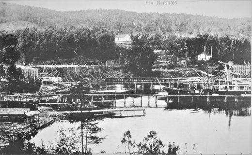 Nærsnesbukta med isstabler og isrenner 1900-1903. En båt som laster is. Forløperen til Propsebrygga er den nærmeste brygga på bildet. Det er isrenne fra isstabel på Mellomstranda. I tillegg er det to isstabler på Flatstranda og i vannkanten ved Bryggebakken. På teglverkets område ser vi tørkehus til venstre i bildet. Den nærmeste brygga går ut fra Nærsnesstranden. Dampskipet ligger ved issklia fra Mellomstranda. Bak den er den lange isbrygga ved Mellomstranda. Nærsnes kapell med takrytter i bakgrunnen. (Foto fra Røyken Historielag, fotograf ukjent)