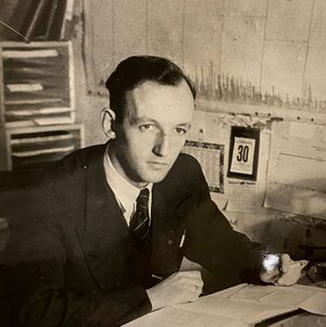 Mann med mørkt hår som sitter ved pult. Penn i høyre hånd. Ser til venstre