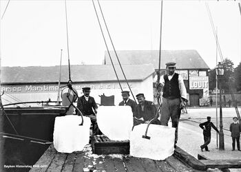 D/S KROSSFOND LOSSER NÆRSNESIS I EUROPEISK HAVN ca. 1900 .Is fra losses fra D/S Krossfond i Saint-Nazaire i Frankrike. To isblokker («gross blocks») henger i issaksene på hver side av vekta. Isblokka som er på vekta har vært stuet inntil skutesida, og er derfor avrundet. Mannskap og de som noterer vektene på isen står i bakkant. Bak sees et av lagerhusene til et av de transatlantiske franske rederiene Messageries maritimes Marseille, som hadde fast havn også i havner ved Atlanterhavskysten/Den Engelske kanal, (Foto Røyken Historielag, fotograf ukjent)