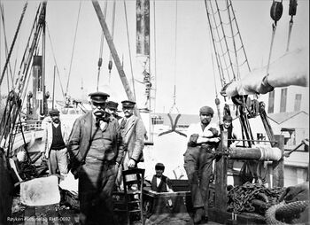 S/S DANNEBROG LOSSER NÆRSNESIS EUROPEISK HAVN ca 1900 Mannskap oppstilt for fotografen på Dekket av S/S Danebrog ved lossing av Nærsnesis. En isblokk henger i issaksa over lasteluka, en er på vekta og en er på dekket. En av mannskapet står i lastrommet, og en står ved vinsjen. Stolen på dekket brukes av en av de som noterer vekta på isen. Vekta er skjult bak skipperen, som står nærmest. I bakgrunnen sees skorsteinen på et dampskip og lagerhus.(Foto Røyken Historielag, fotograf ukjent)