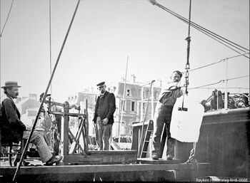 NÆRSNESIS LOSSES I EUROPEISK HAVN.En isblokk henger i issaksa på en dampbåt for å bli plassert på vekta som står på dekket. Bildet er est sannsynlig fra Saint-Nazaire i Frankrike, tatt samtidig med et annet bilde ved lossing av is fra D/S Krossfond. Til venstre sitter iseksportør Adam Baarsrud. (Foto Røyken Historielag, fotograf ukjent)