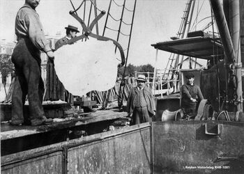 NÆRSNESIS LOSSES I EUROPEISK HAVN.Lossing av is fra Nærsnes i Europeisk havn. En isblokk henger i issaksa for å bli satt ned på vekta til venstre i bildet. Bak isblokka, på brygga, står det stabler med props. Props kunne ofte være dekkslast når hovedlasta var is. Bak den hengende isblokka står Iseksportør Adam Baarsrud. (Foto Røyken Historielag, fotograf ukjent)