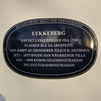 Blå oval byhistorisk plakett på lys vegg i Fredrikstad. Skiltet forteller om Lykkeberg, et stedsnavn kjent fra 1780, kjøpt av grosserer Julius N. Jacobsen i 1855, med nåværende villa oppført 1873–1875, senere brukt til kommune- og kulturadministrasjon.