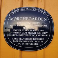 Blå oval byhistorisk plakett på gul trevegg i Fredrikstad. Skiltet forteller om Mørchegården, forstadens eldste kjøpmannsgård, bygget etter brannen i 1812 av skipper Lars Mørch, og fødestedet til Arne Stangebye