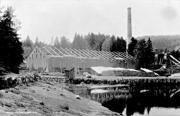 Fra byggingen av teglverket på Nærsnes i 1899. Bygningene på gården Nærsnesstranden ble revet for å gi plass til Teglverket. Bygningen som er under oppførelse, er huset med ovnene. Ovnene og pipa er ferdi bygget. På bildet kan du se de ferdige ovnene synlige inne i ovnshuset. Ovene er Noen lave tørkehus ble plassert mellom ovnshuset og vannet. Til venstre ser veien som gikk fra Nærsnesstranden videre ut til Nærsnes. De samme steinen støtter opp kyststien som går her i dag. Mellom veien og byggeplassen renner Sokkabekken ut. Konstruksjonen helt til høyre i bildet er en del av isstabelen på Mellomstranda. (Foto 1899 fra Røyken Historielag, fotograf ukjent)'