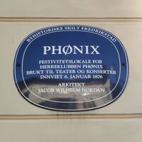 Blå oval byhistorisk plakett på lys vegg i Fredrikstad. Skiltet forteller om Phønix, et festlokale for herreklubben Phønix, brukt til teater og konserter, innviet i 1876 og tegnet av arkitekt Jacob Wilhelm Nordan