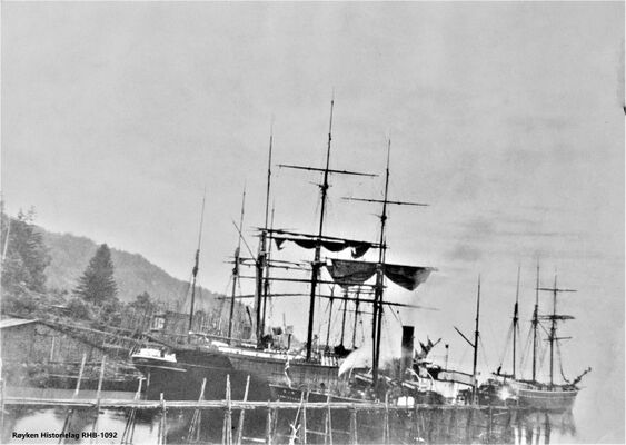 FIRE SEILSKUTER OG ET DAMPSKIP LASTER IS. 1890-1900 Ved den lange isrenna (nærmest) ligger D/S Ran av Kristiania. En isblokk er på isrenna ved båten. Bak skipene ligger den store isstabelen på Bryggebakken. I venstre kant av bildet ligger en stor stabel props klar til lasting. I tillegg til is ble det eksportert props fra Nærsnes. Props kunne være dekkslast når isen var hovedlast. (Foto fra Røyken Historielag)