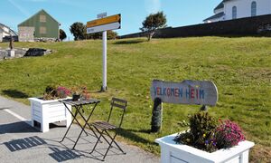 Velkomsthelsing på ferjekaien. Statuen av fiskarkona til venstre. Løa til bnr 19 Stuberg bakom, og kyrkja bak til høgre. Bilde 2021.