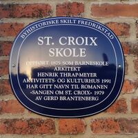 Blå oval byhistorisk plakett på murvegg i Fredrikstad. Skiltet forteller om St. Croix skole, oppført i 1875 som barneskole, tegnet av arkitekt Henrik Thrap-Meyer, og senere brukt som aktivitets- og kulturhus