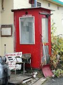 Telefonkiosk i tre på verftstomta til Kaarbøs Mek. Verksted, Harstad, i samme farge som originalen og en kopi av Fastings originale kiosk, bygd på dugnad av verftsarbeiderne for å slippe å gå opp på et av kontorene for å ringe ut eller motta beskjeder. Foto: Gunnar E. Kristiansen (2016).