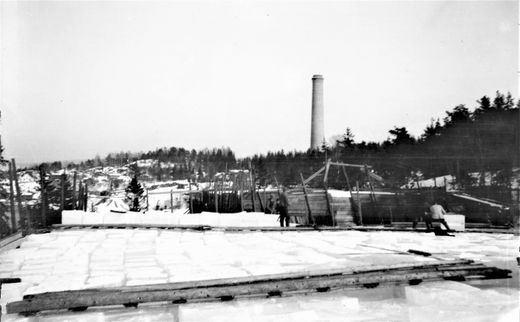 TOPPEN AV ISSTABEL PÅ FLATSTRANDA 1900-1915. I forgrunnen ligger ledere for flytting av isblokker. Til høyre hales en isblokk ut på en leder. Teglverkspipa er i bakgrunnen. Bildet tatt før topp-tildekking med sagflis som isolasjon. (Foto fra Jon Hovind)