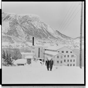 To menn går opp fra den hvite fabrikke med skortesteine som ses i bakgrunne