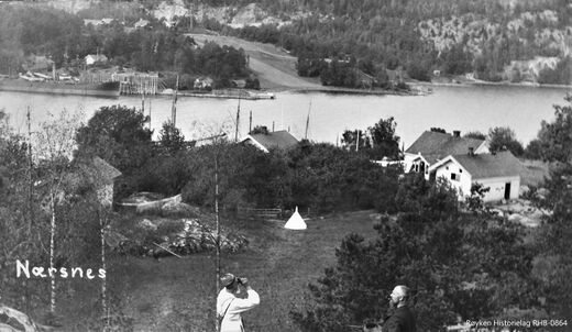 UTSIKT OVER BUKTA 1905-1912.Fra området over/bak Lundesteinen mot Graffstranda. En stor lastebåt laster is ved den lille isbrygga rett innenfor isstabelen i vannkanten av Bryggebakken. Isstabel på Graffodden med isrenne over jordene inn fra Scwarzedammen. Bak kapellet (nå Nærsnes Kirke) som har takrytter – ikke spir, ser vi Søndre Nærsnes med låven. En stor lastebåt laster is ved Bryggebakken. Huset med to piper i forgrunnen er kjøpmann Lundes første butikklokaler (Foto fra Røyken Historielag, fotograf ukjent)